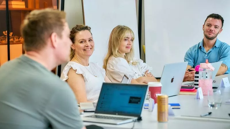 Group of colleagues sitting around a table with laptops and drinks, engaged in a relaxed meeting and smiling.