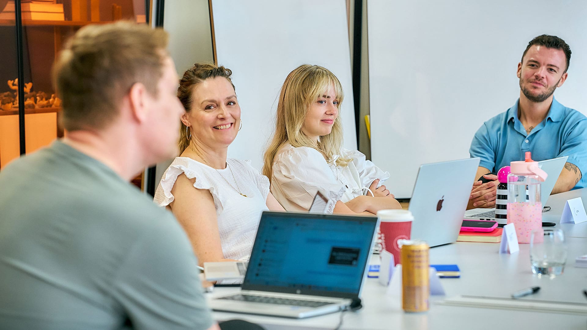 Group of colleagues sitting around a table with laptops and drinks, engaged in a relaxed meeting and smiling.