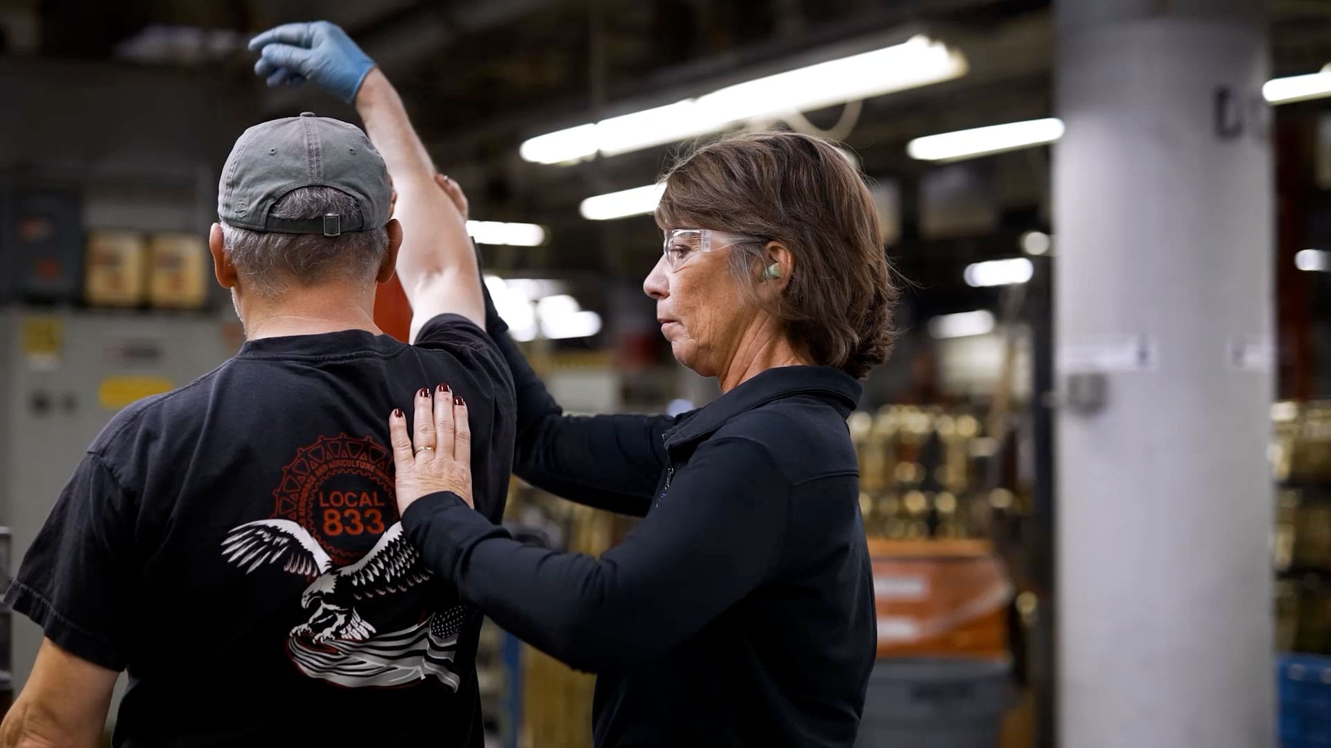 A woman in safety glasses assists a man in a factory by guiding his raised arm, both wearing protective gear in an industrial setting.