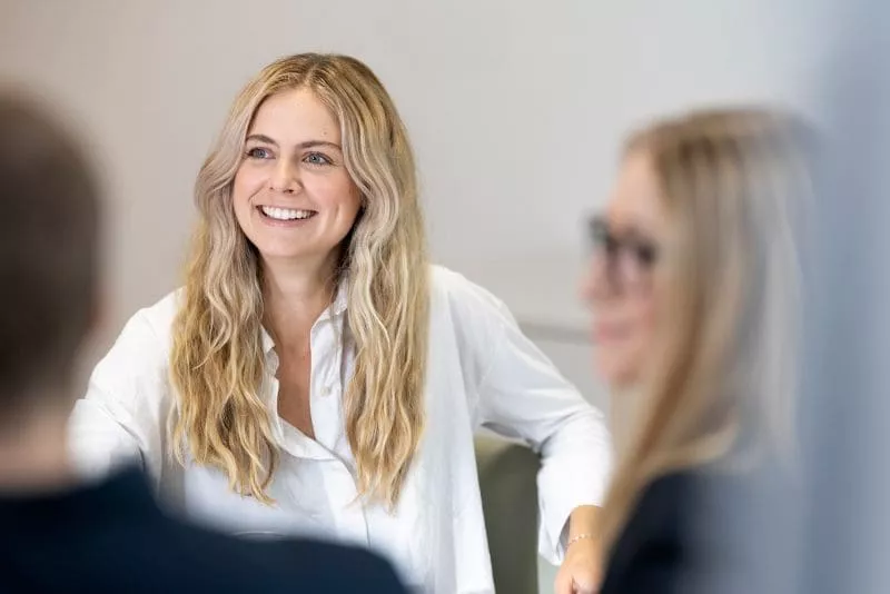 Woman with long blonde hair smiling during a meeting in a bright office setting.