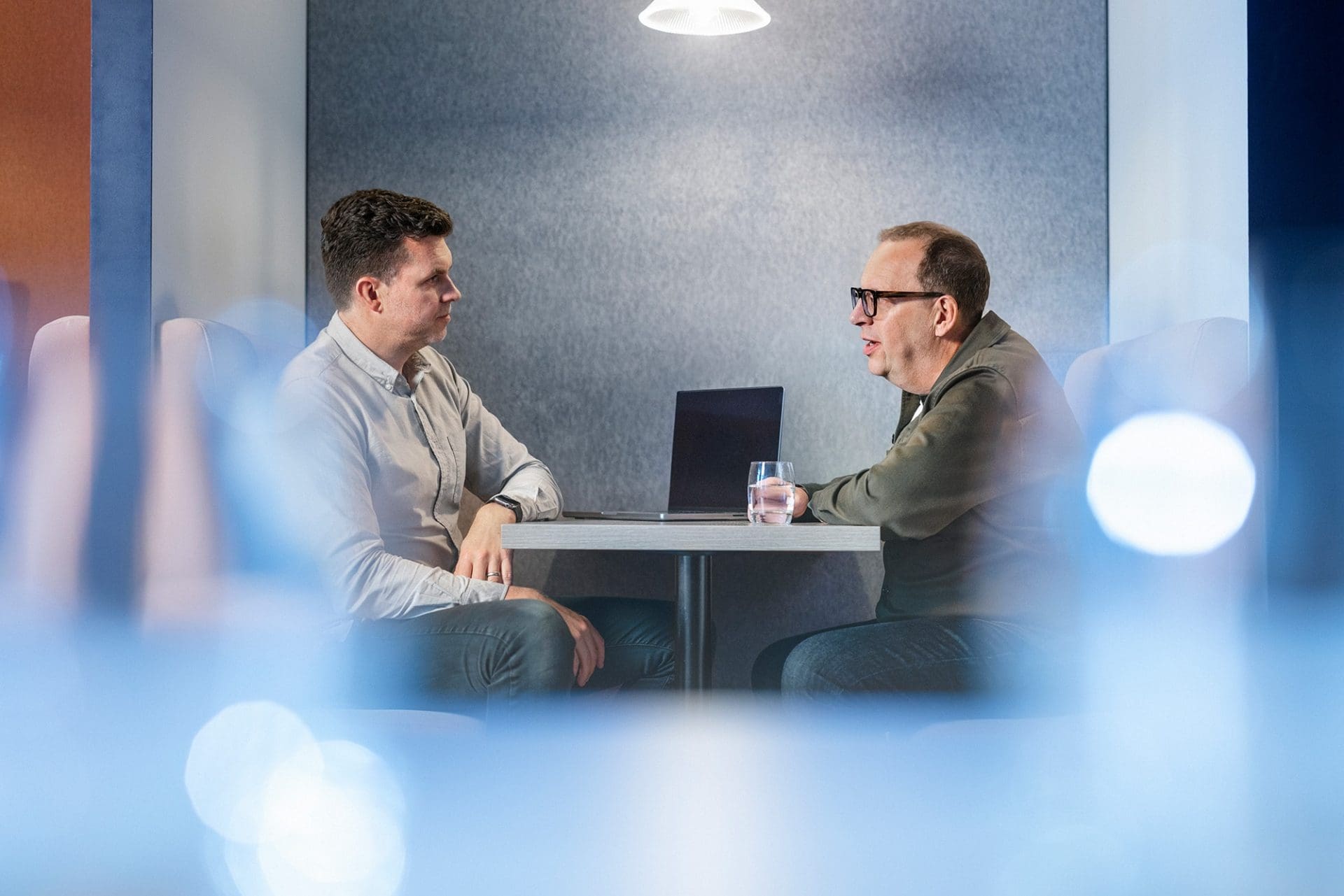 Two men having a discussion at a small table in a modern office booth with a laptop and a glass of water.