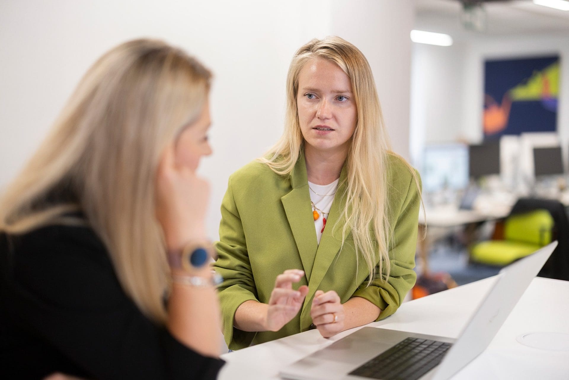Two people in discussion at a desk with a laptop in a modern office setting.