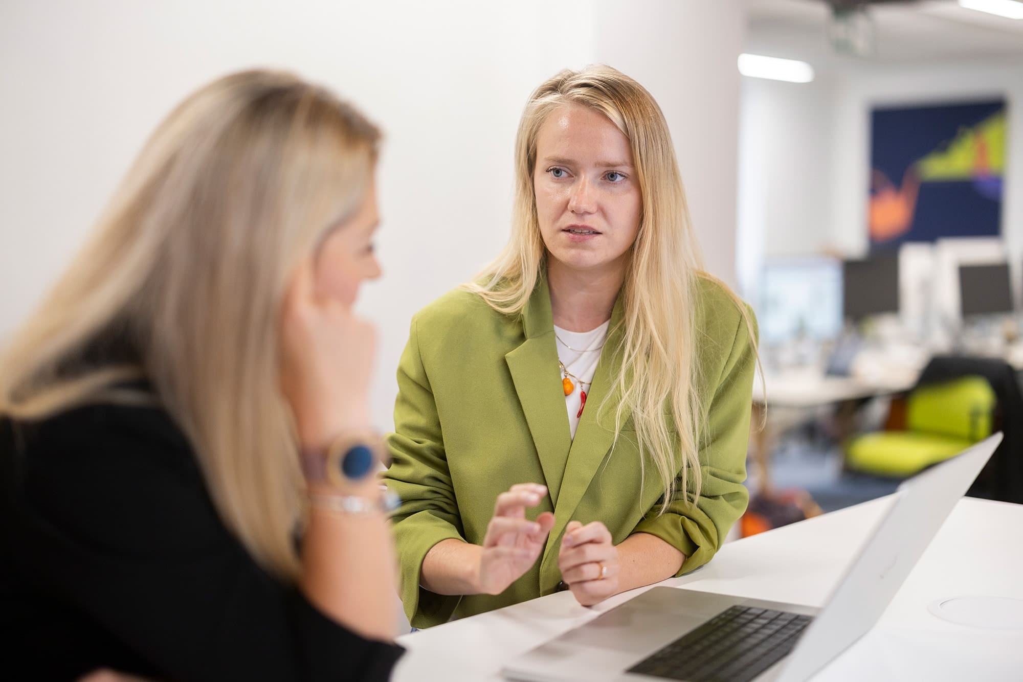 Two people in discussion at a desk with a laptop in a modern office setting.