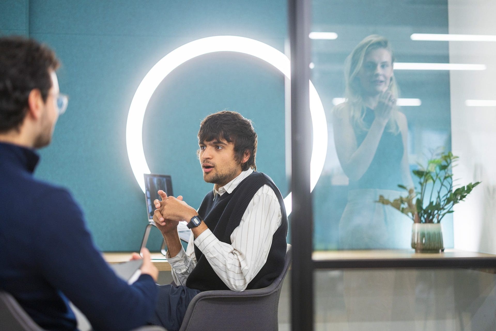 Two people having a discussion in an office meeting room, with a bright circular light on the wall behind them.