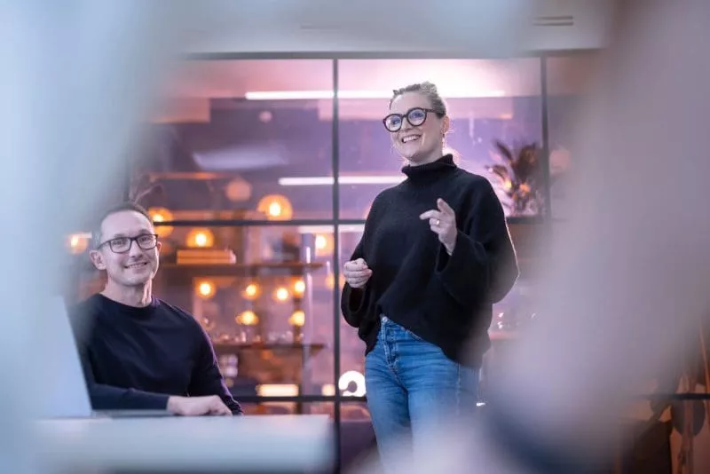 Woman standing and speaking with a smile during a meeting, while a man seated nearby listens, in a modern office with warm lighting and glass walls.