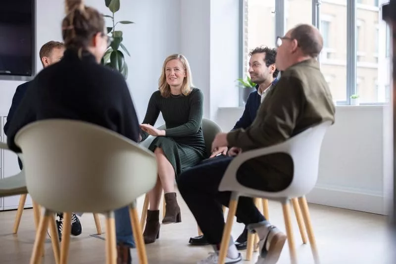 Group of five people sitting in a bright modern office, with one woman speaking while the others listen and smile.