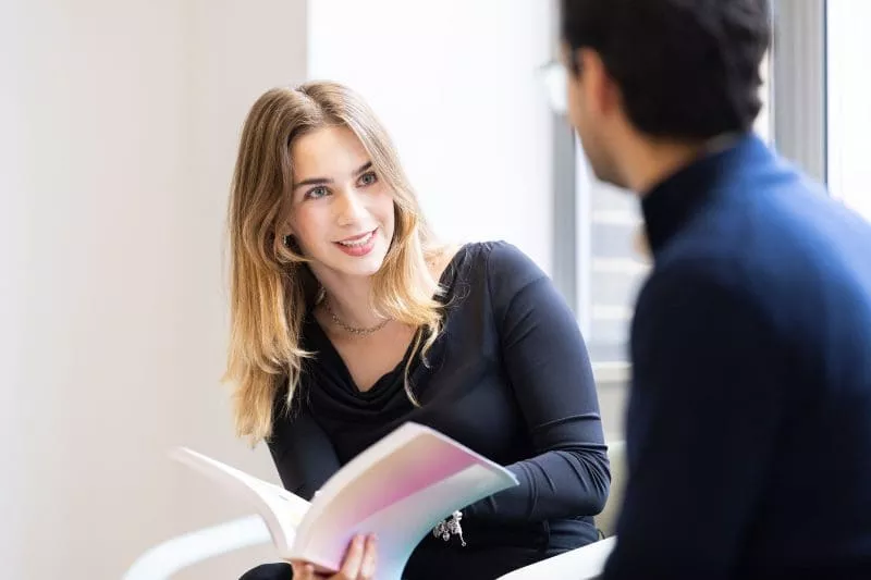 Lady reading book and talking to colleague in office setting.