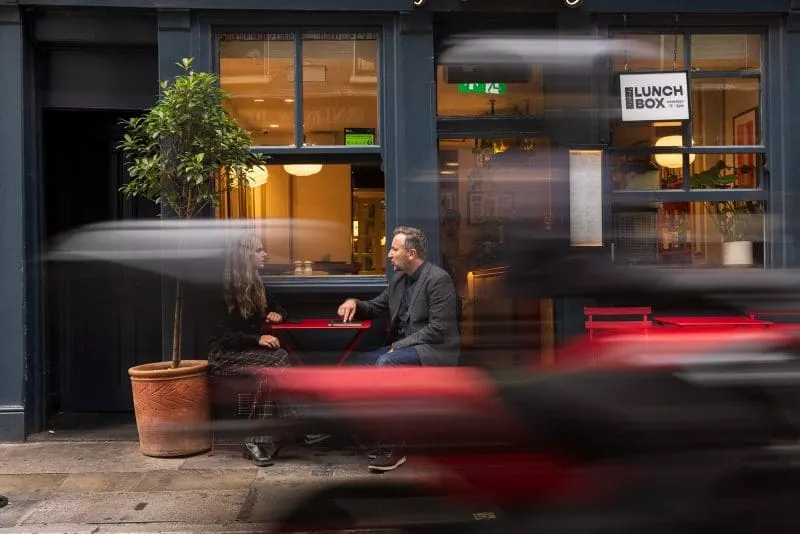 Two people sitting at a small table outside a café, with a motorbike passing by in motion blur.