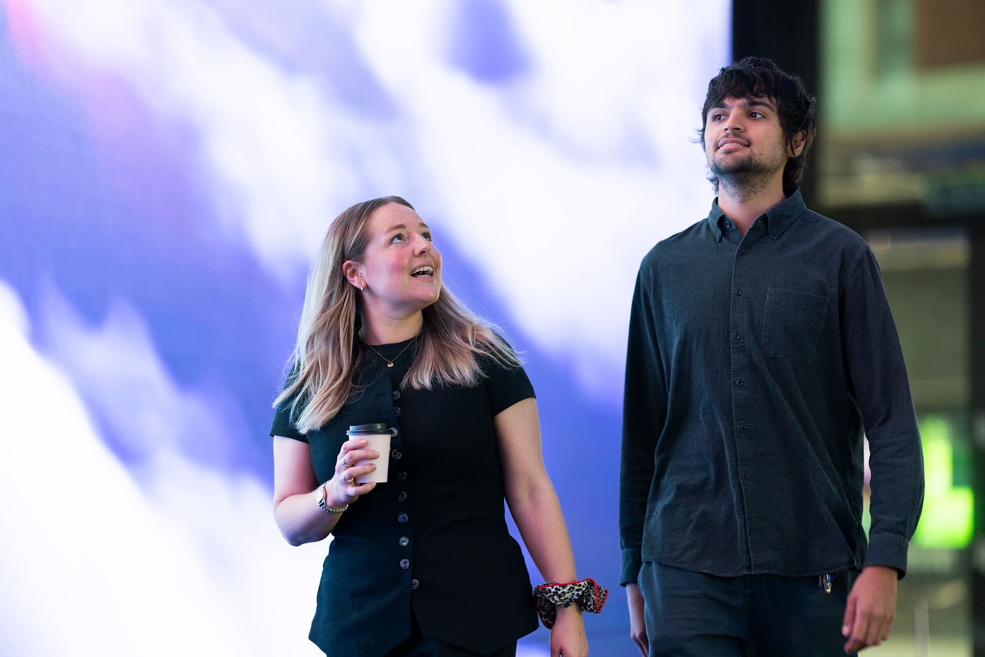 Two people walking and talking in front of a large illuminated screen with abstract blue and white light.