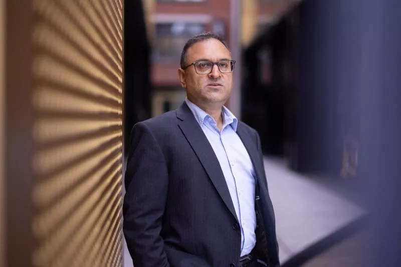 Person wearing glasses and a suit standing outdoors beside a patterned golden wall.