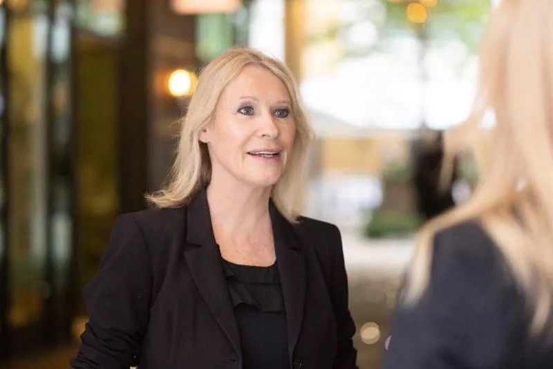 Person wearing a black jacket speaking to someone indoors with soft lighting and a blurred background.