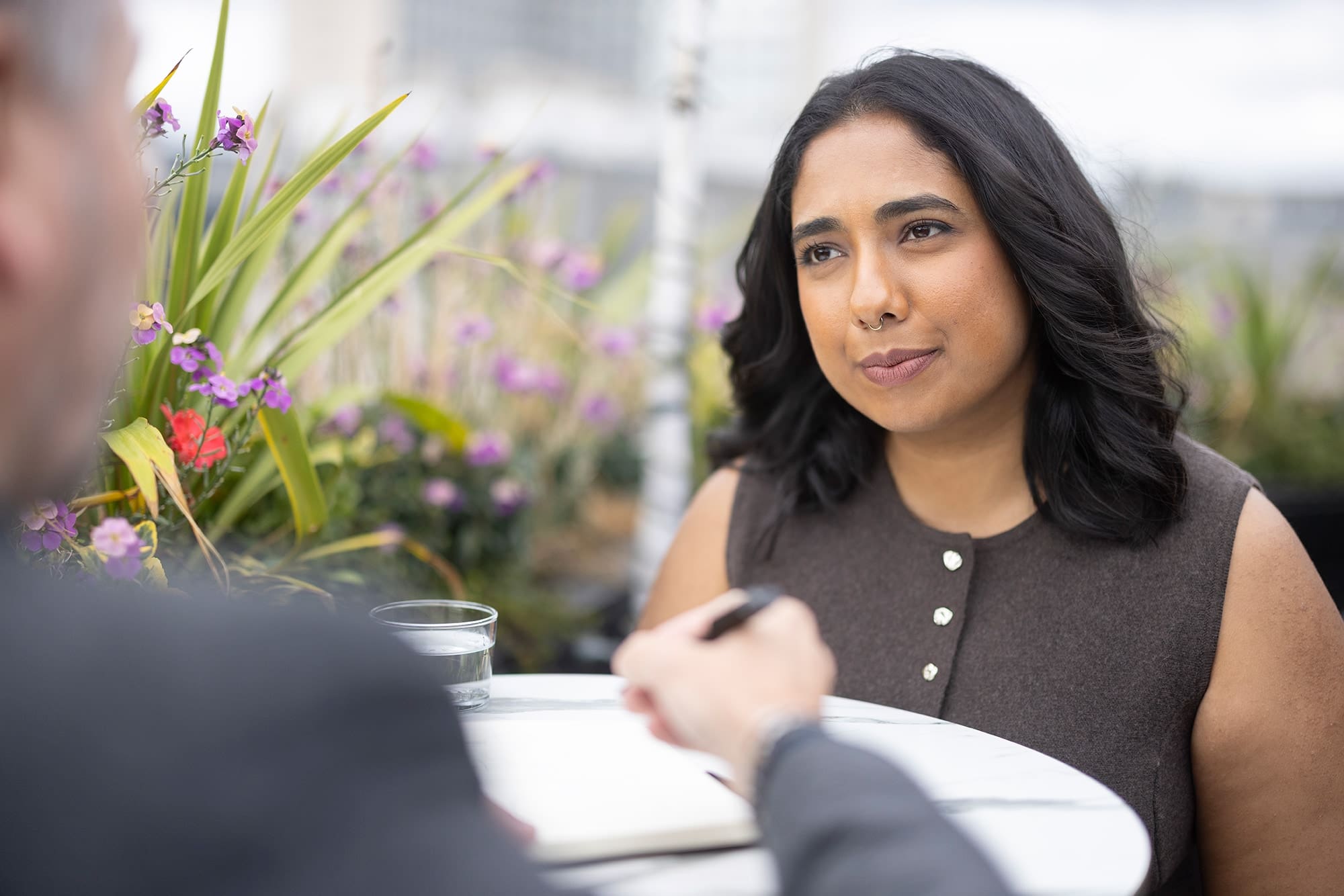 Woman sitting at an outdoor table with flowers in the background, listening attentively to someone during a conversation.