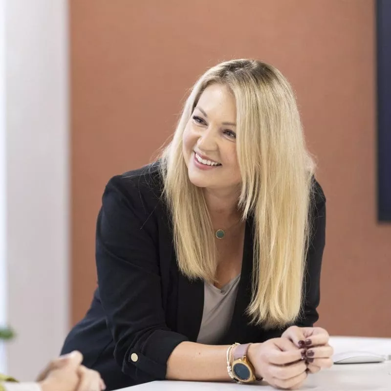 Person smiling and leaning on a table during a conversation, wearing a black blazer.