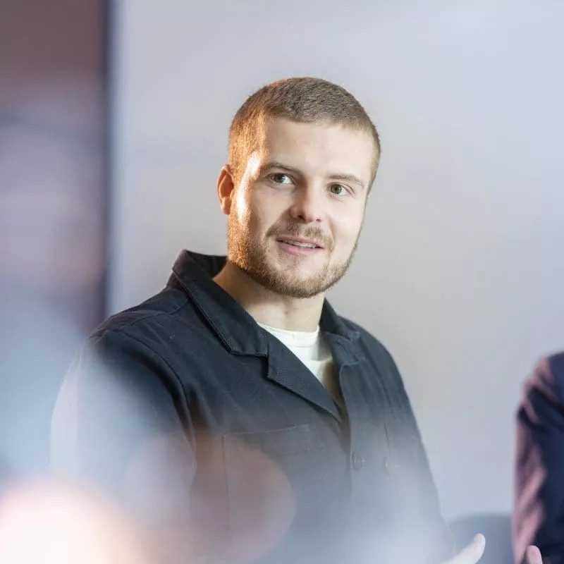 Person speaking in a meeting, wearing a dark jacket and gesturing with hands.