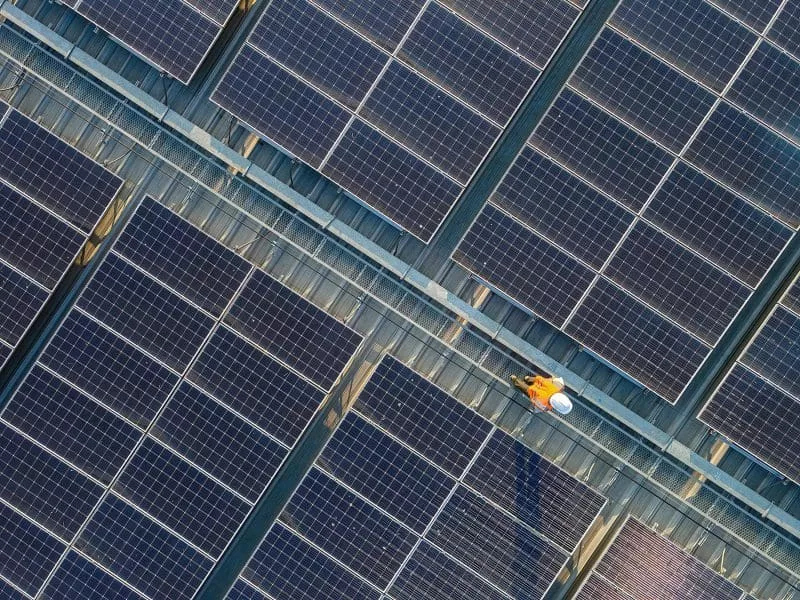 Aerial view of a worker in safety gear inspecting a large rooftop solar panel array.