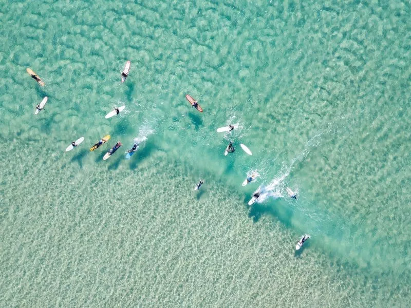 Aerial view of surfers paddling in clear turquoise ocean water over sandy seabed.