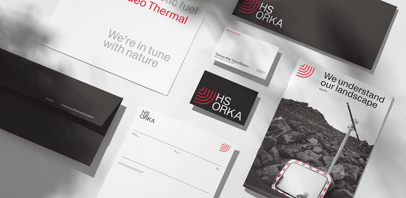 A flat-lay photograph of branded stationery and print materials from HS Orka’s visual identity. At the top right is a dark folder with the red and white HS Orka logo. Below and to the left are business cards in black and white with the logo and a person’s name. In the center are white sheets of paper and envelopes with minimal printed lines and the HS Orka wordmark. At bottom right is a promotional brochure or booklet cover showing a rugged rocky landscape, a curved red graphic accent, and the phrase “We understand our landscape” alongside the HS Orka logo. All elements are arranged neatly on a light surface, showcasing the brand’s clean typography and use of red, black, and white in its identity design