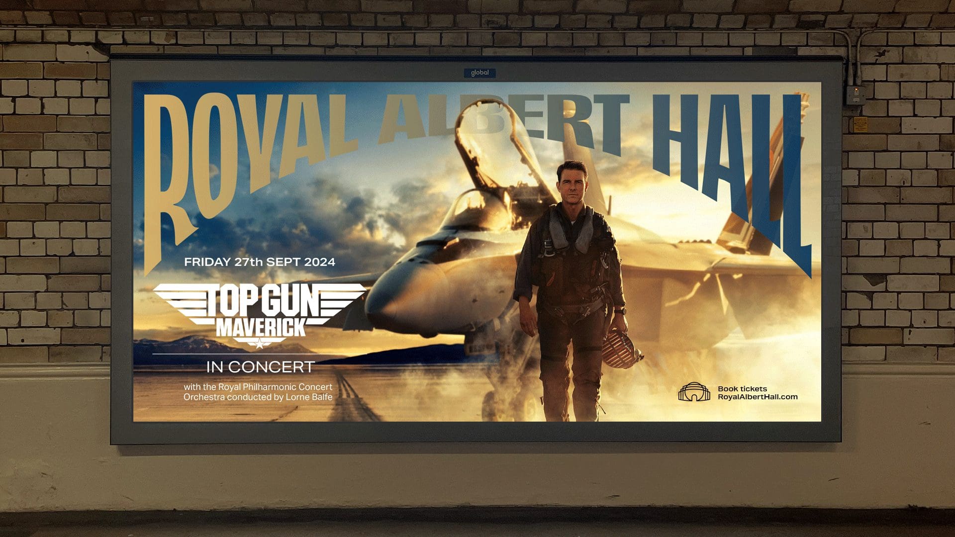 A large landscape advertising poster displayed on a brick wall in a transit or outdoor setting. The poster shows a dramatic scene from Top Gun: Maverick with a pilot in flight gear standing in front of a fighter jet against a cloudy sky. Above him, large stylised letters spell out “ROYAL ALBERT HALL” in a curved masthead.