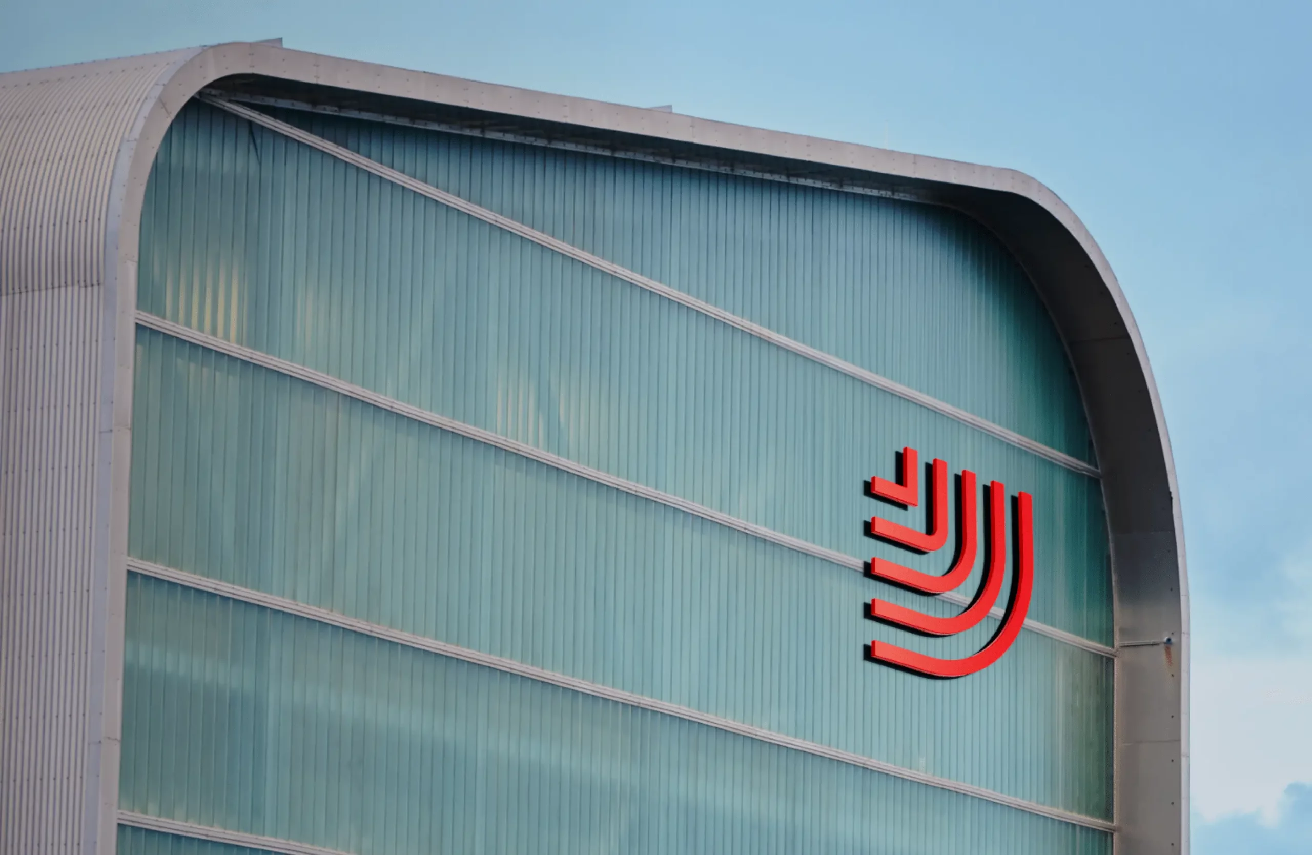 A close-up of the side of a modern industrial building with curved rooflines and large vertical glass or metal panels. On the greenish-blue façade is a bold red graphic logo made of several curved lines forming a stylised abstract emblem. The sky above is clear with soft light illuminating the structure. (Image from HS Orka brand project by Brandpie showing branded signage on a building exterior.)
