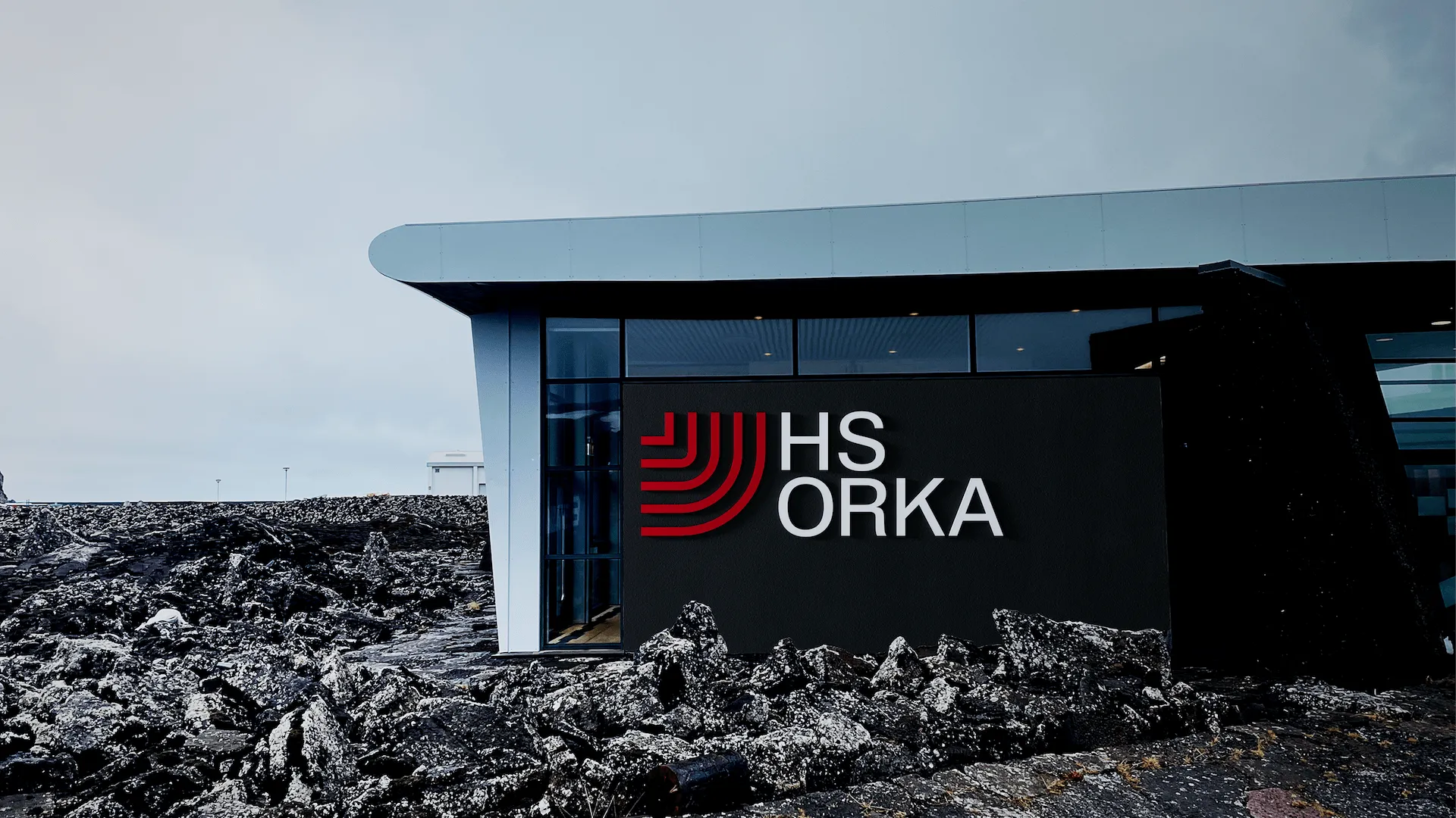 A photograph of the exterior of a modern industrial building in an open landscape, showing part of HS Orka’s facility with clean architectural lines and branded signage. The structure has a large flat façade with the red HS Orka logo or graphic mark prominently displayed against a light-colored wall, and the sky above is clear or lightly clouded. The composition highlights the building’s scale and the brand’s presence on the site, reflecting the company’s role in renewable energy production in Iceland.